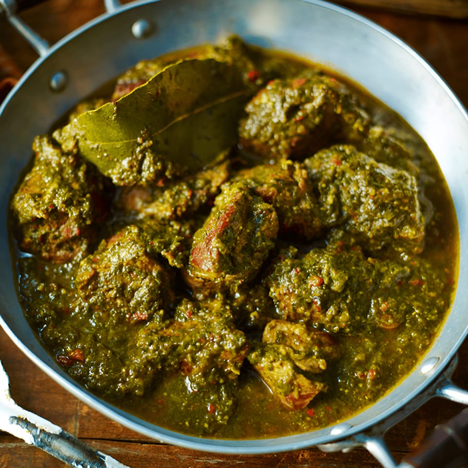 Virasat Indian Cuisine's green Indian curry with meat pieces and a bay leaf in a metal pan.